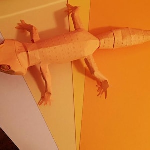 以下が含まれることがあります： オレンジ色の紙でできたトカゲのペーパークラフトモデル。トカゲは足を伸ばして、尻尾を丸めてポーズをとっています。