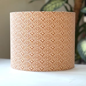 May include: Cylindrical lampshade featuring a repeating geometric pattern in tan and cream. The pattern is made up of diamond shapes, giving a textured look. The lampshade is designed to fit over a lamp base and provide ambient lighting.