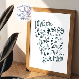 Op de afbeelding: Een ingelijst print met een witte achtergrond en een handgeschreven citaat in blauw en grijs: "LOVE the Lord your God with all your heart & with all your soul & with all your mind"  Mattheüs 22:37