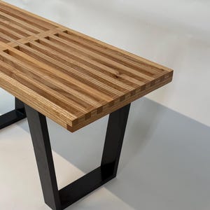 May include: A bench featuring a slatted seat made of light brown wood with visible grain patterns. The seat is supported by two black metal legs with a rectangular frame design. The slats are arranged horizontally with small gaps between them.