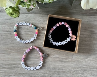 May include: Three colourful beaded bracelets with the word "SCHULKIND" spelled out on each. The bracelets are in a pink, blue and green colour scheme. The bracelets are in a box with a black background.