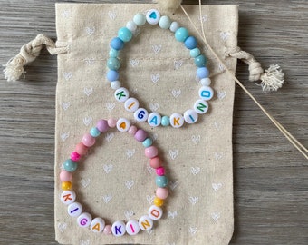 Könnte beinhalten: Zwei Perlenarmbänder mit dem Wort "KIND" in bunten Buchstaben. Die Armbänder liegen auf einer beigefarbenen Stofftasche mit einem weißen Herzmuster.