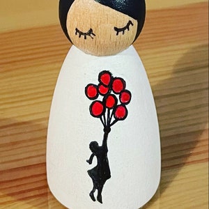 以下が含まれることがあります： 黒い髪と白いドレスを着た木製のドール。ドールは赤い風船を持っており、ドレスには女の子の黒いシルエットが描かれている。