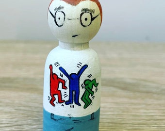 May include: A hand-painted wooden peg doll with brown hair, glasses, and a white shirt with a colorful abstract design. The doll is wearing blue pants.