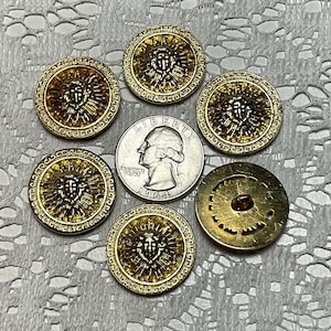 May include: Six gold-toned buttons with a raised design of a face with a crown and a sunburst pattern. The buttons have a Greek key border and are approximately 1 inch in diameter.