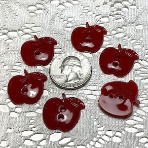 May include: Six red plastic apple-shaped buttons with two holes each. The buttons are arranged on a white lace background with a US quarter for scale.