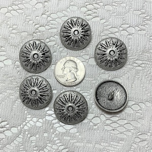 Peut inclure: Six boutons en métal argenté avec un design texturé en relief. Les boutons sont de forme circulaire avec un centre surélevé et un dessus légèrement bombé.