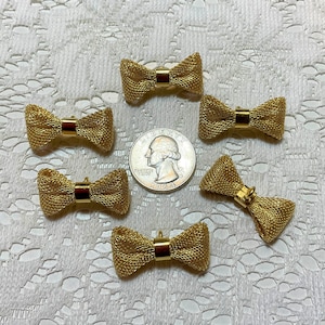 May include: Six gold-colored metal bow tie charms with a textured finish. The charms are arranged on a white lace surface.