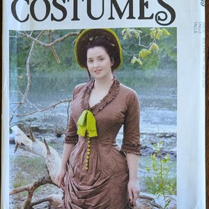 May include: McCall's Costumes sewing pattern featuring a woman in a brown dress with a green bow and buttons. The pattern cover includes the text "McCall's COSTUMES" and "Angela Clayton". The woman is wearing a hat with a green brim.
