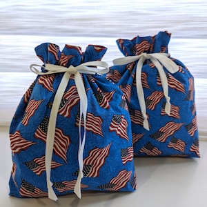 May include: Two blue drawstring bags with white ribbon ties. The bags are decorated with a pattern of red, white, and blue American flags.