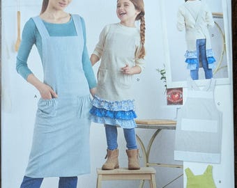 May include: Simplicity sewing pattern S9436 featuring aprons. The image shows a woman and a child wearing aprons, with additional apron designs. The pattern includes sizes XS-XL. The text "Designed and printed in USA" is visible.