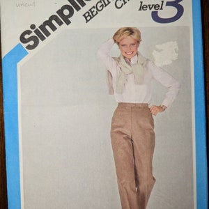 May include: Simplicity Beginner's Choice sewing pattern, level 3, size 18, waist 32 inches. The pattern features a woman wearing a light pink shirt and brown pants. The text "Shirt & sweater not included" is visible.