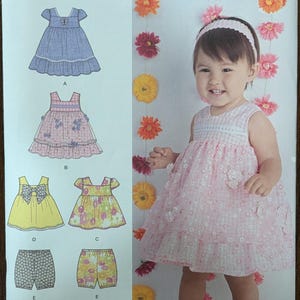 May include: Simplicity pattern 1471A for baby dresses and shorts. The pattern includes several dress designs, including a blue dress with a butterfly detail and a pink floral dress. The image also shows a baby wearing a pink dress with floral accents.