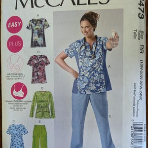May include: McCall's M6473 sewing pattern envelope for scrub tops and pants. The front displays illustrations of various scrub top styles with floral and abstract prints, alongside solid-colored pants. Text includes Easy, Plus, and size range 18W-24W.