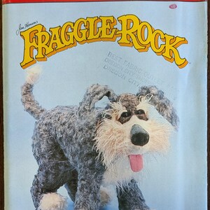 May include: A vintage Butterick craft pattern featuring a plush Fraggle Rock dog. The pattern cover has the text "FRAGGLE ROCK" in yellow, with the Butterick logo and pattern details. The dog is gray, white, and pink.