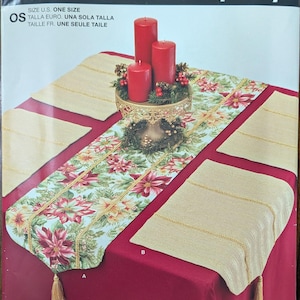 May include: A Simplicity pattern for a table runner and placemats. The table runner has a floral design with red and cream flowers. The placemats are beige with gold trim. Red candles and a gold candle holder are also visible.