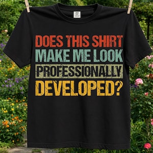 May include: A black short-sleeved t-shirt featuring distressed, multi-coloured text that reads: DOES THIS SHIRT MAKE ME LOOK PROFESSIONALLY DEVELOPED? The lettering is arranged in four horizontal rows with red, light blue, dark green, and yellow colours.