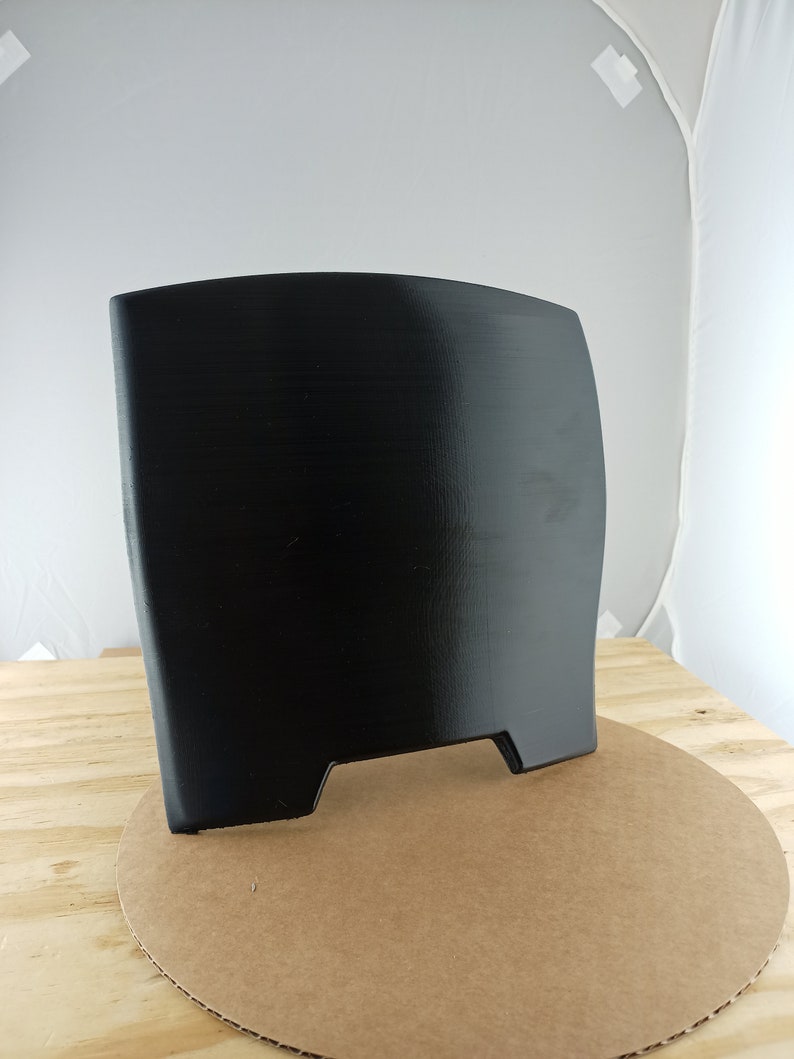 May include: A black plastic shield with a curved top and two small cutouts at the bottom. The shield is standing on a brown cardboard circle.