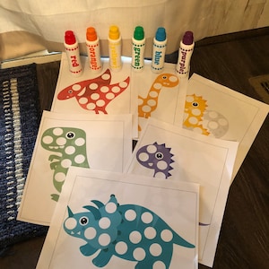 May include: A set of six dinosaur dot-to-dot activity sheets with corresponding color markers. The markers are labeled with the colors red, orange, yellow, green, blue, and purple.