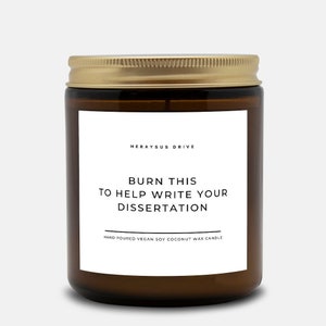 May include: A brown glass candle jar with a gold lid. The label reads "HERAYSUS DRIVE" and "BURN THIS TO HELP WRITE YOUR DISSERTATION". The candle is hand-poured vegan soy coconut wax.
