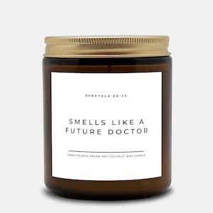 May include: A brown glass candle jar with a gold lid. The label reads "Herayus Drive" and "Smells Like A Future Doctor" with the text "Hand Poured Vegan Soy Coconut Wax Candle" below.