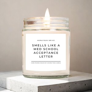May include: A clear glass jar candle with a white label that reads "HERAYSUS DRIVE SMELLS LIKE A MED SCHOOL ACCEPTANCE LETTER HAND POURED VEGAN SOY COCONUT WAX CANDLE". The candle is lit and the flame is visible.