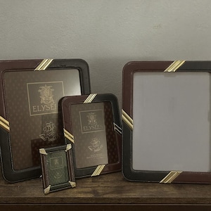 May include: A collection of leather picture frames in various sizes. The frames are brown and black with gold accents and the word "ELYSEES" printed on the inside. The frames are displayed on a wooden surface.