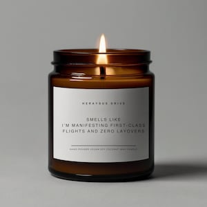 May include: A brown glass candle jar with a white label that reads "HERAYSUS DRIVE" and "SMELLS LIKE I'M MANIFESTING FIRST-CLASS FLIGHTS AND ZERO LAYOVERS". The candle is lit and the wax is melting.