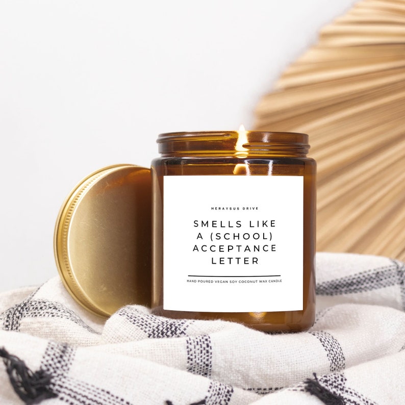 May include: A brown glass candle with a gold lid. The candle is lit and has a white label that reads "Smells like a (school) acceptance letter". The label also includes the text "Herayus Drive" and "Hand poured vegan soy coconut wax candle".