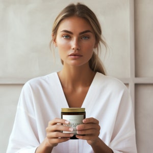 May include: A woman in a white robe holds a candle with a gold lid. The candle label reads "Smells Like A Nap Day".