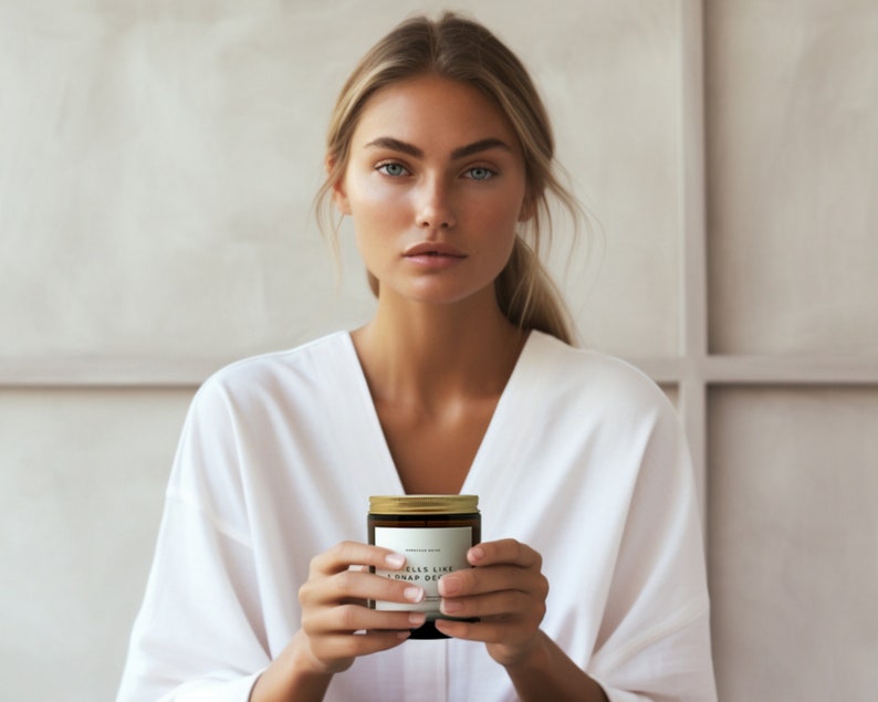 May include: A woman in a white robe holds a candle with a gold lid. The candle label reads "Smells Like A Nap Day".