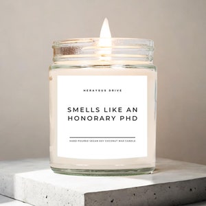 May include: A clear glass jar candle with a white label that reads "Herayus Drive" and "Smells Like An Honorary PhD" with the text "Hand Poured Vegan Soy Coconut Wax Candle" below. The candle is lit and the flame is visible.