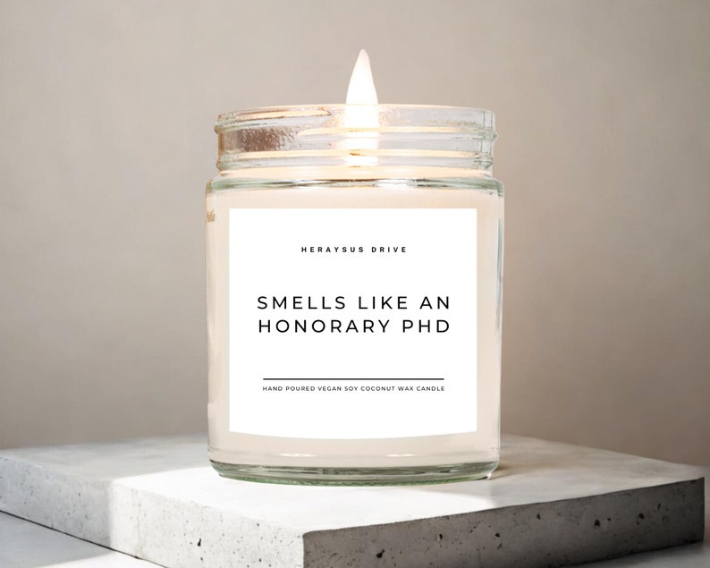 May include: A clear glass jar candle with a white label that reads "Herayus Drive" and "Smells Like An Honorary PhD" with the text "Hand Poured Vegan Soy Coconut Wax Candle" below. The candle is lit and the flame is visible.