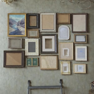 May include: A collection of various vintage picture frames in different shapes, sizes, and styles, arranged on a textured gray wall. The frames are made of wood and metal, with some containing artwork or blank spaces.