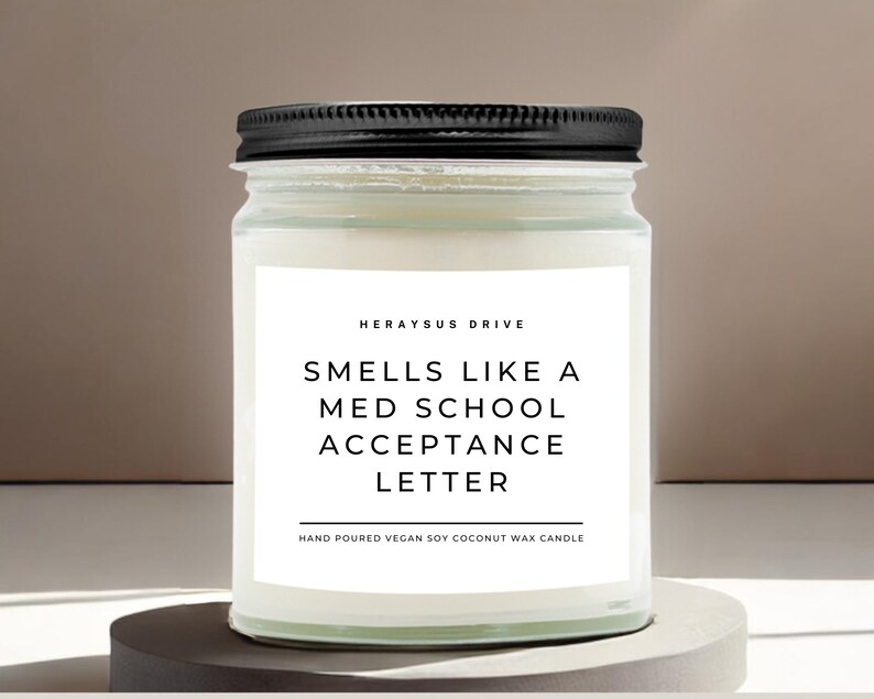 May include: A white candle in a glass jar with a black lid. The label reads "HERAYSUS DRIVE SMELLS LIKE A MED SCHOOL ACCEPTANCE LETTER HAND POURED VEGAN SOY COCONUT WAX CANDLE".