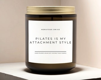May include: A brown glass candle with a gold lid and a white label. The label reads "Herayus Drive" and "Pilates is my attachment style". The label also says "Hand poured vegan soy coconut wax candle".