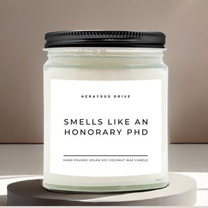May include: A white candle in a clear glass jar with a black lid. The label reads "Herayus Drive" and "Smells Like An Honorary PhD". The label also says "Hand Poured Vegan Soy Coconut Wax Candle".