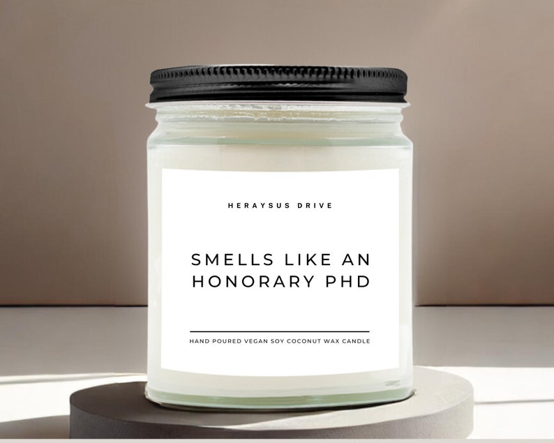 May include: A white candle in a clear glass jar with a black lid. The label reads "Herayus Drive" and "Smells Like An Honorary PhD". The label also says "Hand Poured Vegan Soy Coconut Wax Candle".