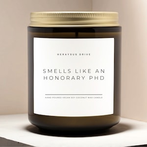 May include: A brown glass candle jar with a gold lid. The label reads "Herayus Drive" and "Smells Like An Honorary PhD" with the text "Hand Poured Vegan Soy Coconut Wax Candle" below.