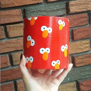 Peut inclure: Un pot cylindrique rouge avec un motif répété d'yeux blancs et de nez orange. Le pot est peint pour ressembler à Elmo de Sesame Street.