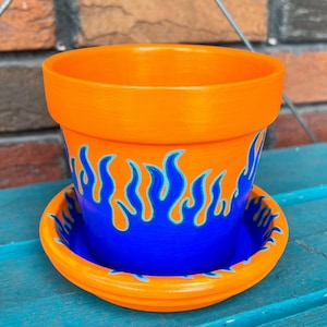 Peut inclure: Pot en terre cuite orange avec un motif de flammes bleues et turquoise. Le pot a une soucoupe assortie.