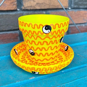 May include: A yellow ceramic planter with a wavy orange design and black and white yin yang symbols. The planter has a matching saucer.