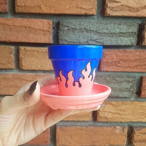 Peut inclure: Un petit pot en céramique bleu avec une soucoupe rose. Le pot est décoré d'un motif de flammes noir et blanc.