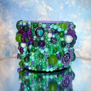 Peut inclure: Un récipient cylindrique orné d'un assortiment de perles vertes, violettes et turquoise. Les perles sont de tailles et de textures variées, certaines translucides. Un petit emblème rond est visible sur le côté.