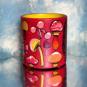 May include: A cylindrical, hand-painted ceramic pot with a vibrant red exterior and a yellow interior. The pot is adorned with colorful illustrations of various mushrooms in shades of red, pink, orange, and yellow. The pot is reflected on a shiny surface.
