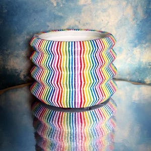 May include: A white ceramic planter with a rainbow striped pattern. The planter has a wavy, textured design and is reflected in a shiny surface.