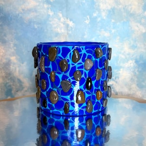 May include: A cylindrical, blue decorative vase with a mosaic pattern. The vase is adorned with various-sized, smooth, dark-colored stones. The background is a light blue with a cloudy pattern, and the vase is reflected on a shiny surface.