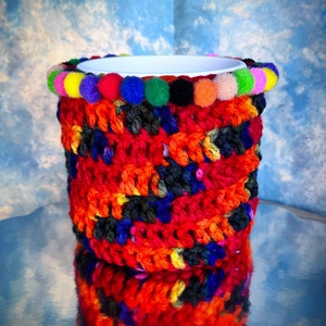 Peut inclure: Un manchon de tasse en crochet avec une bordure de pompons colorés. Le manchon est fait de plusieurs couleurs, dont le rouge, l'orange, le jaune, le vert, le bleu et le noir.
