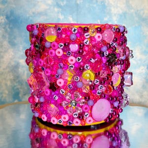 May include: A cylindrical container adorned with pink, purple, and clear beads. The beads vary in size and shape, including round, square, and faceted. Some beads spell out the word "LOVE". The container has a yellow base and rim, reflected in the surface below.