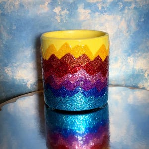 May include: A yellow ceramic pot with a rainbow chevron pattern covered in glitter. The pot is sitting on a reflective surface.